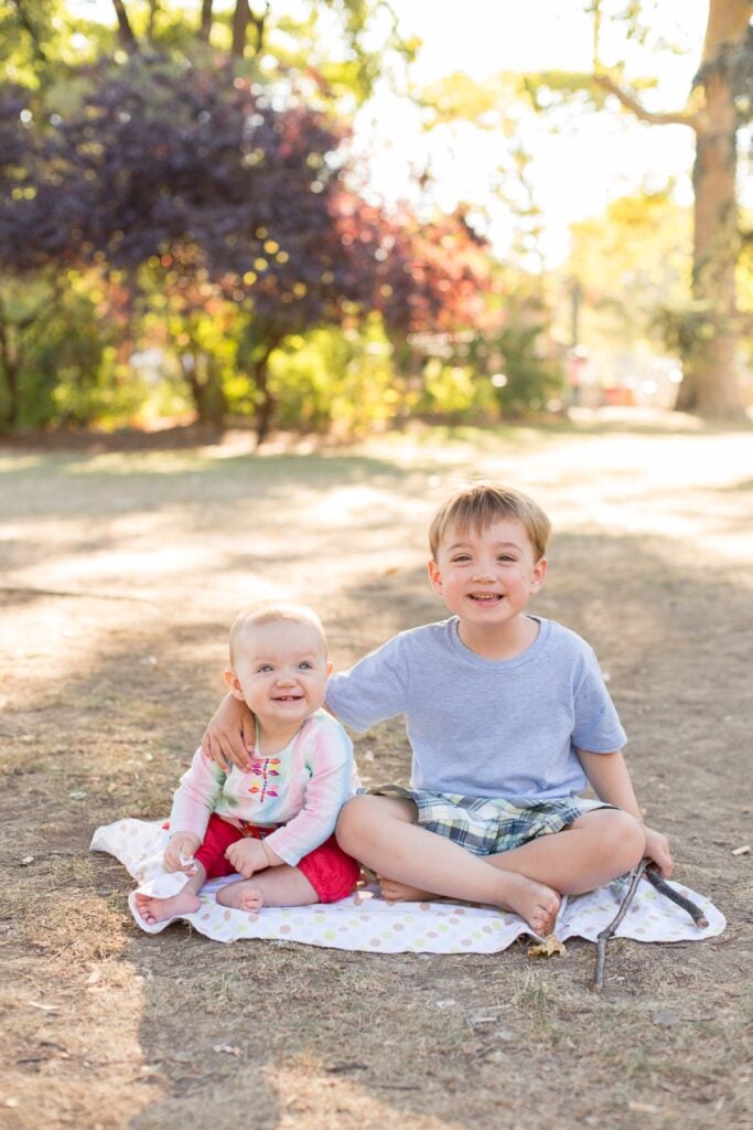 Big brother and baby sister sitting together on a blanket in a Paris park, smiling at the camera
