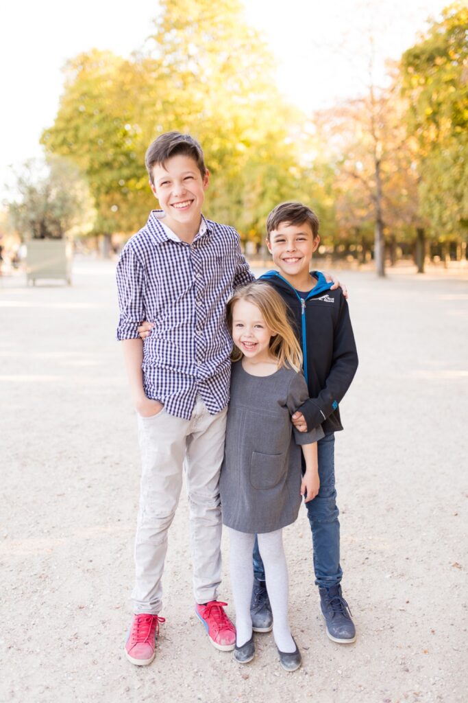 Three siblings hugging and smiling on a tree-lined path in a Paris park
